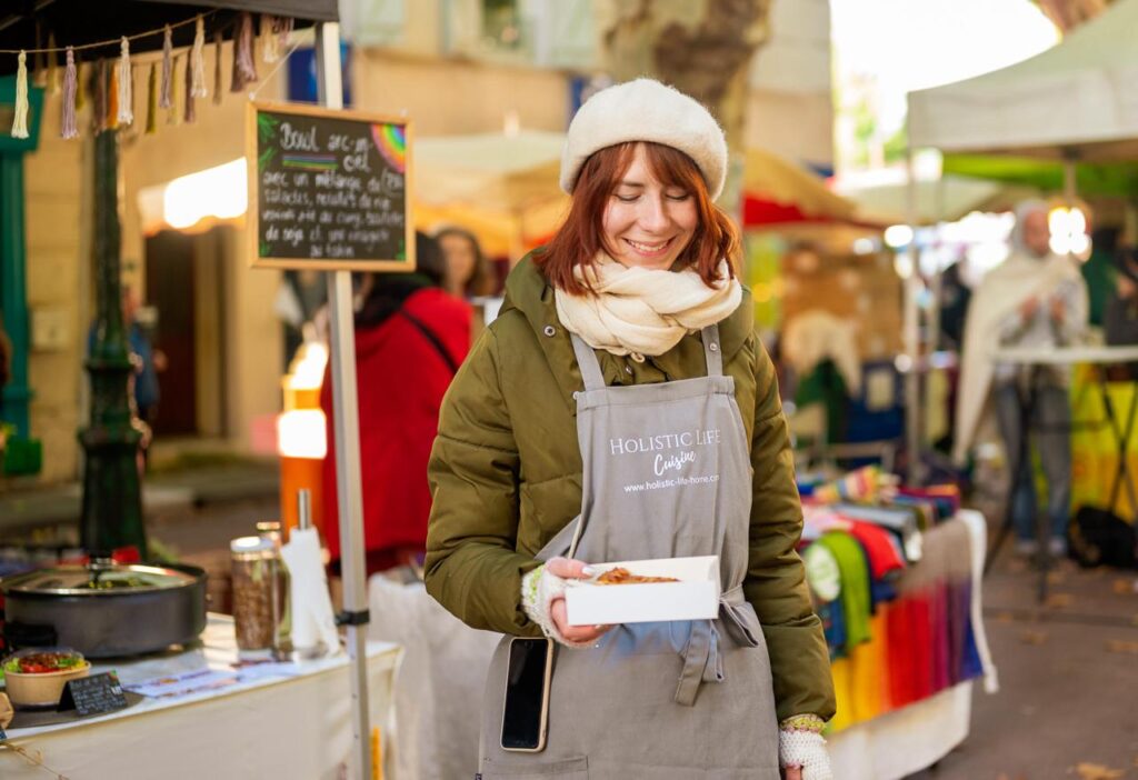 La'inesh steht mit der Schürze, der Holistic Life Cuisine, und der Regenbogen-Bowl in der Hand, vor dem veganen Marktstand.